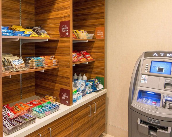 A self-service snack and beverage area with shelves stocked with various snacks and drinks, and an ATM machine to the right.