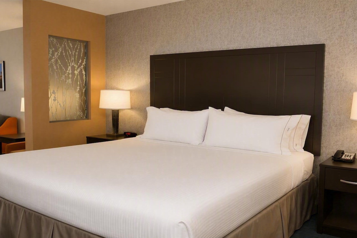 A hotel room with a large bed, two bedside tables with lamps, a nightstand, a chair, and a painting on the wall.