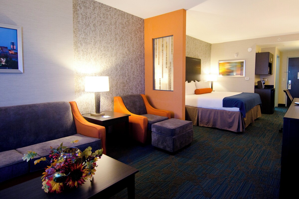 Quarto de hotel com sofá, poltrona, banqueta, cama, escrivaninha e luminária.