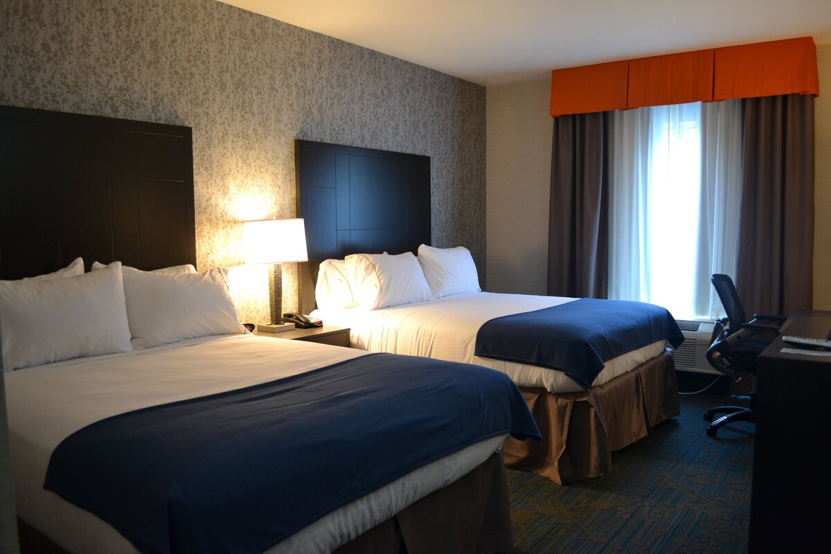 Quarto de hotel com duas camas, uma escrivaninha com cadeira, uma luminária e uma janela com cortinas.