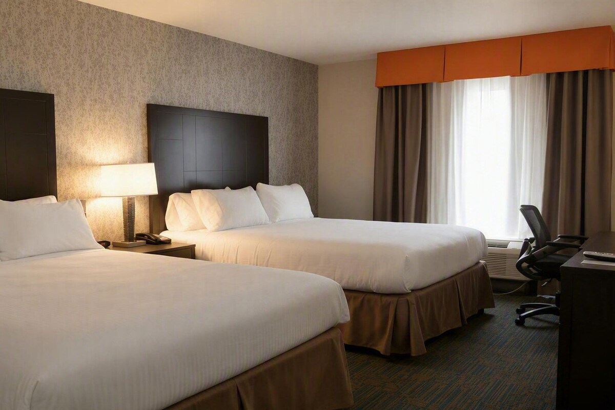 A hotel room with two beds, a desk, a chair, and a large window with curtains.