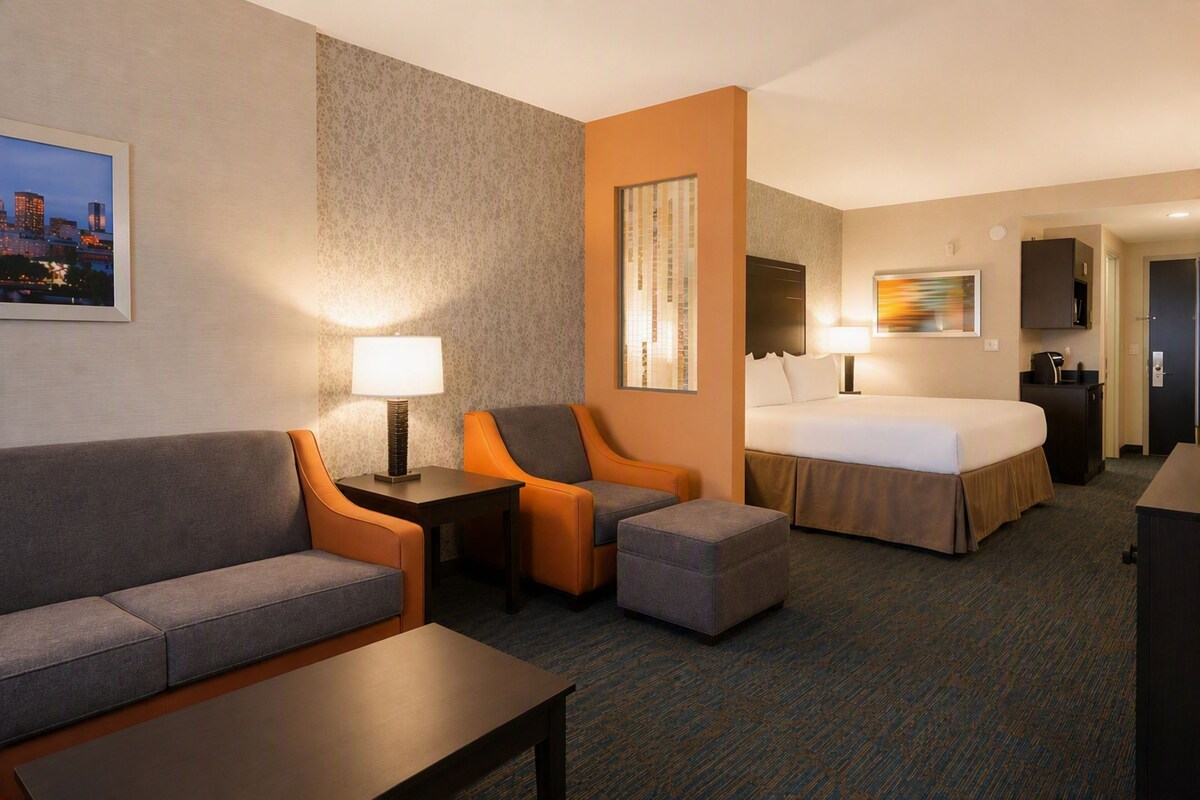 A hotel room with a bed, a sofa, an ottoman, a desk, and a chair. There is a painting on the wall and a lamp on the desk.