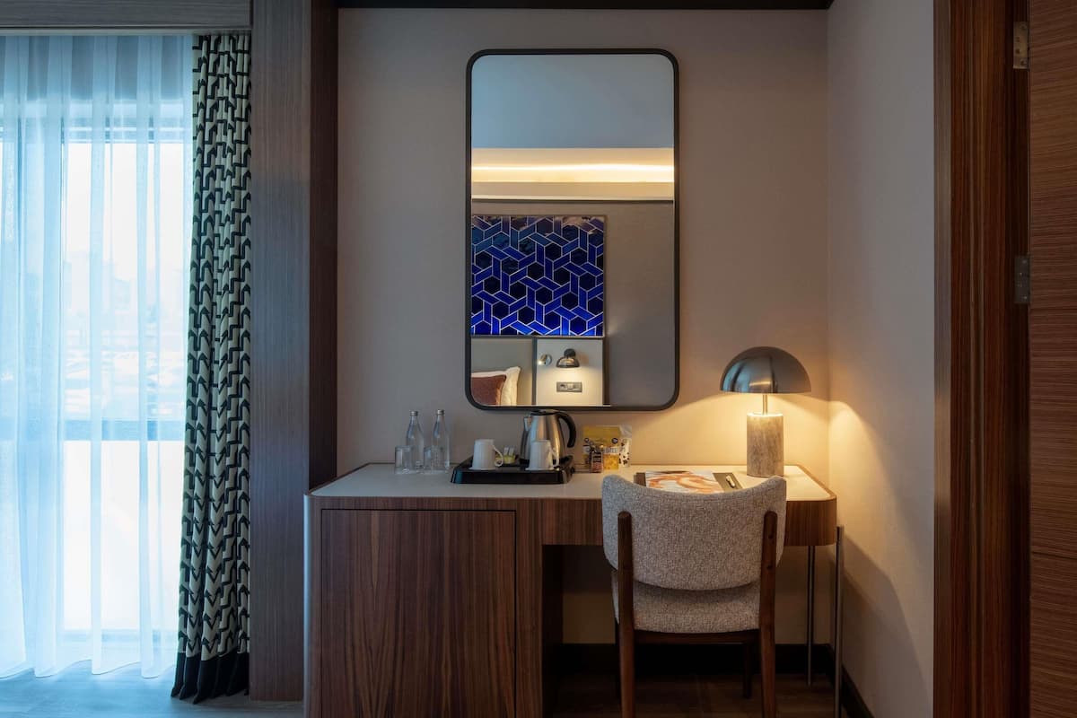 Une chambre dâhĂŽtel avec un bureau en bois, une chaise, un miroir, une lampe et une fenĂȘtre avec des rideaux lĂ©gers.