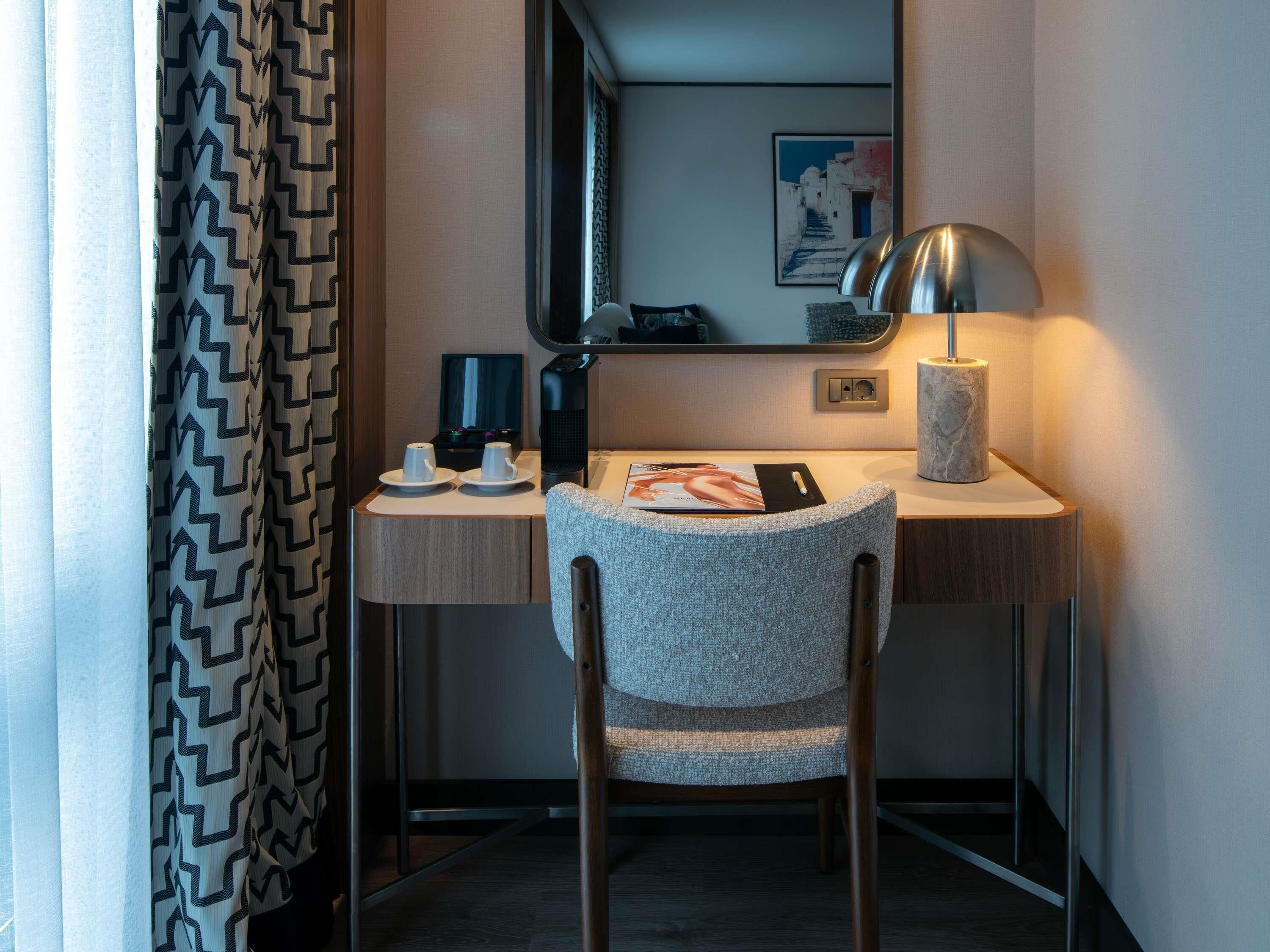A hotel room with a desk, a chair, a lamp, a mirror, and a framed picture on the wall.