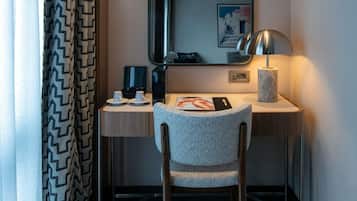 A hotel room with a desk, a chair, a lamp, a mirror, and a framed picture on the wall.