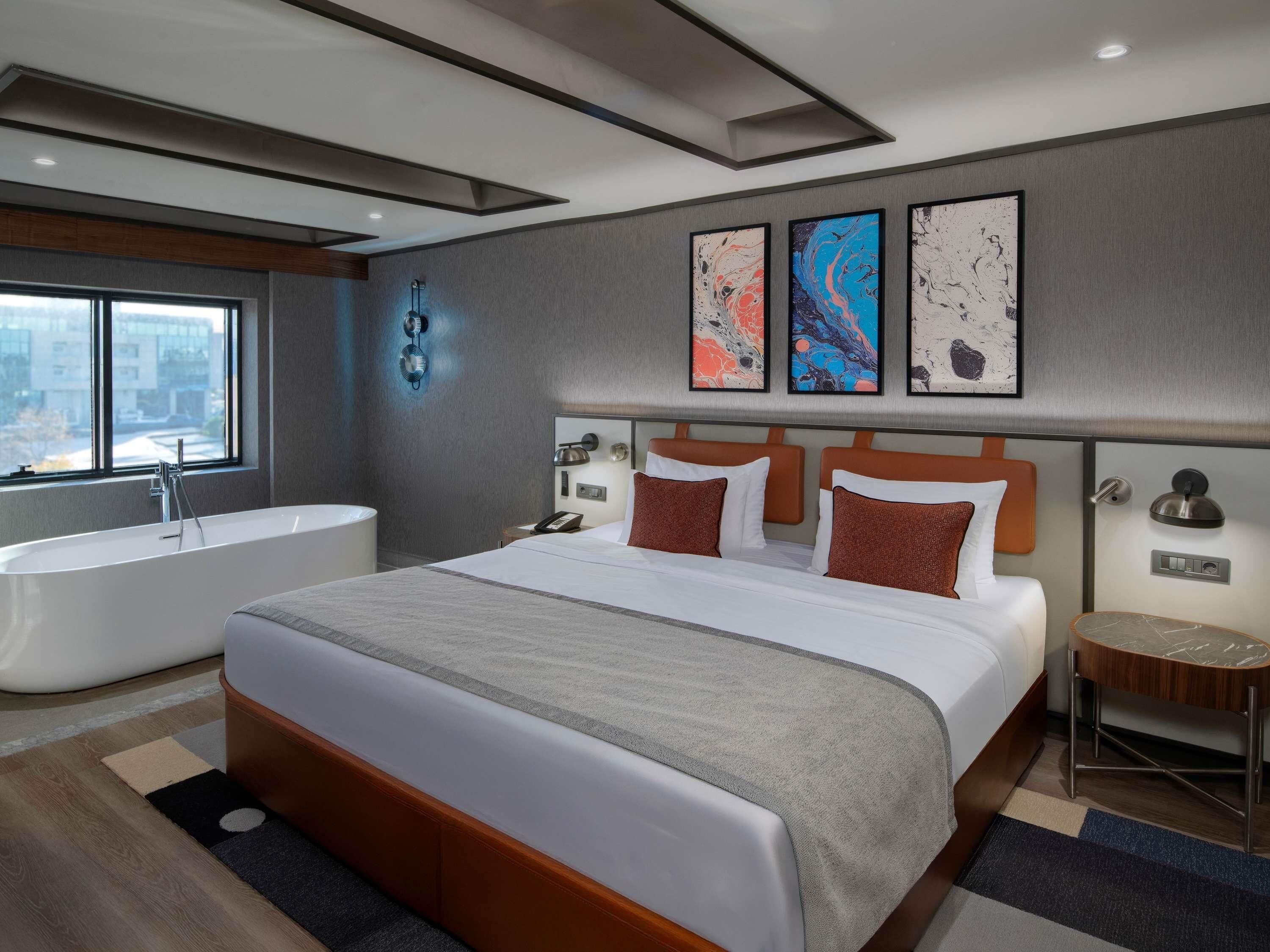 A modern hotel room with a large bed, a separate bathtub, a city view, and abstract art on the wall.