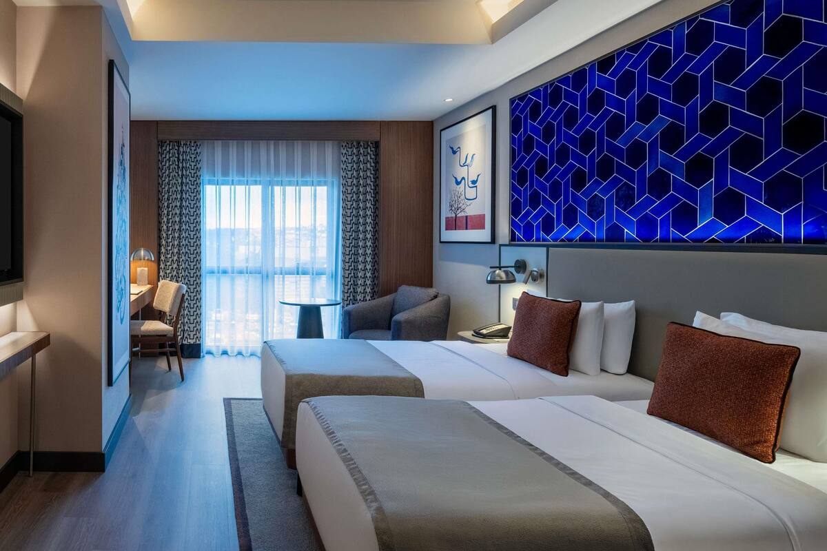 Een hotelkamer met twee bedden, een bureau, een stoel en een groot raam met gordijnen.