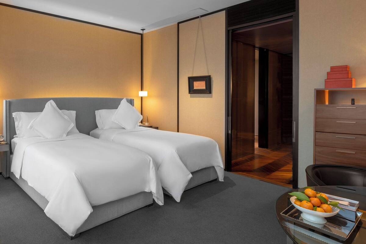 Une chambre d’hôtel avec deux lits, un bureau sur lequel se trouve un panier de fruits et une commode en bois.