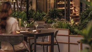 Un área de comedor al aire libre con sillas y mesas de mimbre, sombrillas y un bar visible a través del vidrio.