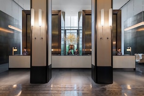 A modern lobby with a reception desk, two elevators, and a large window with a view of greenery.