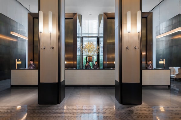 A modern lobby with a reception desk, two elevators, and a large window with a view of greenery.