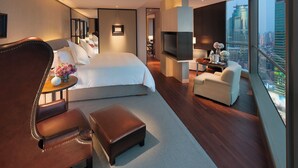 Una habitación de hotel moderna con una cama grande, una zona de estar con una silla y reposapiés, una mesita con flores, un televisor y vistas a la ciudad.