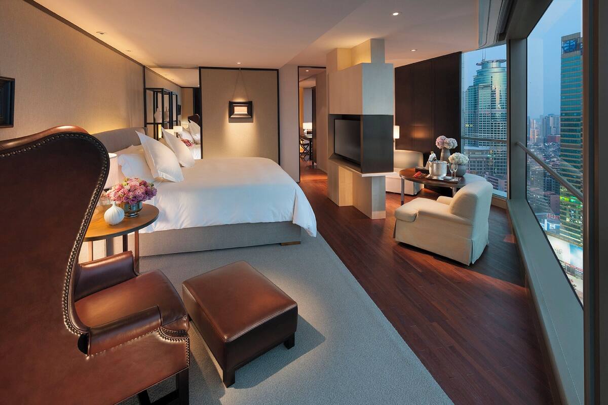 Une chambre d’hôtel moderne dotée d’un grand lit, d’un coin salon avec un fauteuil et un pouf, d’une petite table agrémentée de fleurs, d’une télévision et offrant une vue sur la ville.