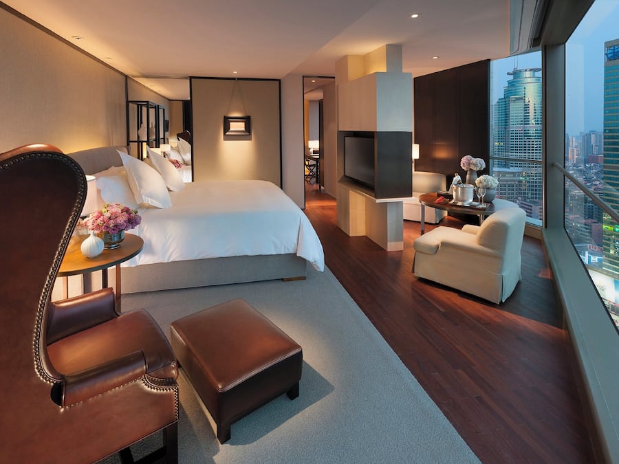 A modern hotel room with a large bed, a seating area with a chair and ottoman, a small table with flowers, a TV, and a city view.