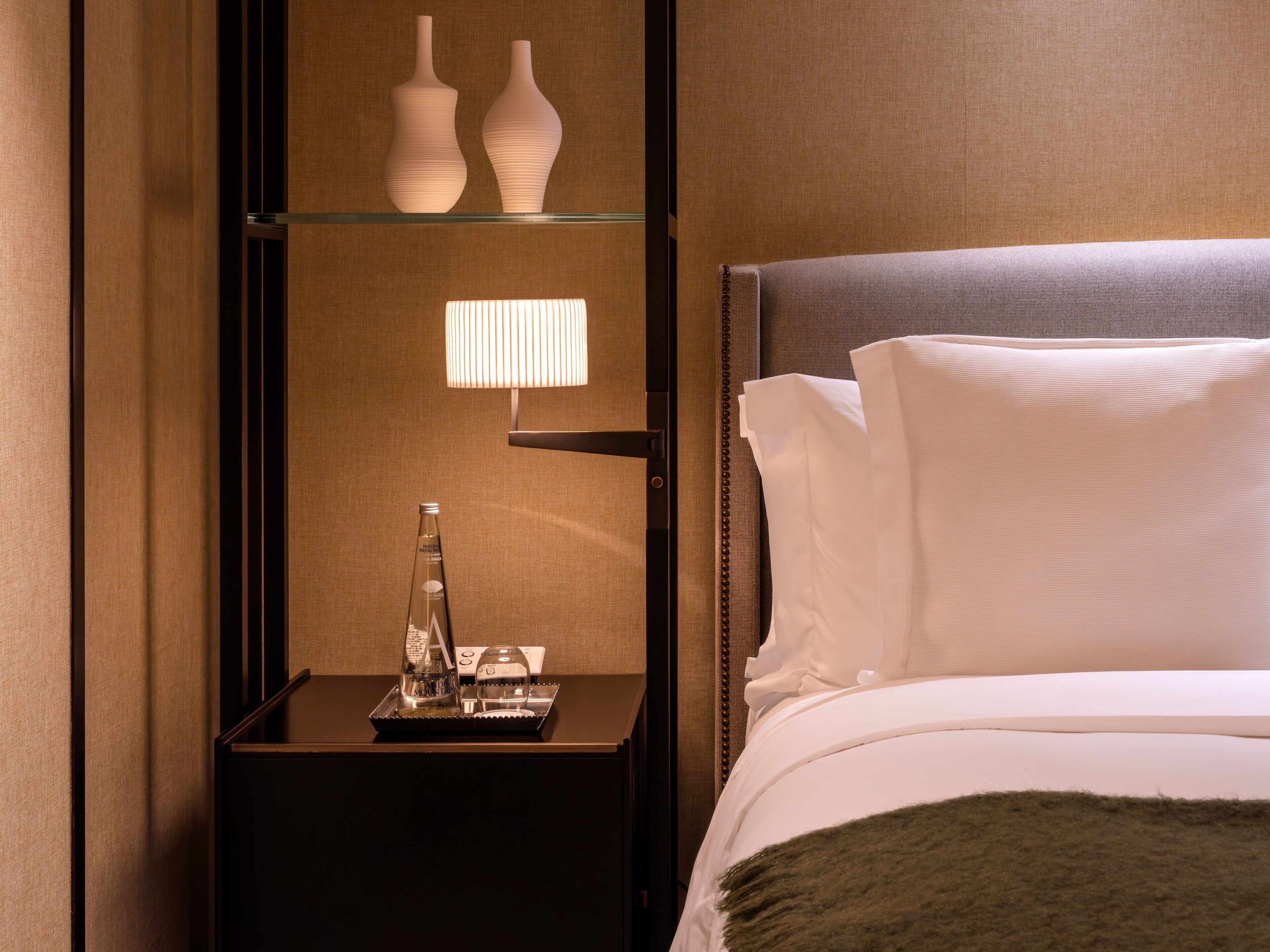 A hotel room with a bed, a nightstand, a lamp, and two vases on the wall.