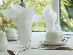 Ein Tisch ist mit weißem Geschirr gedeckt, darunter ein Glas, eine Tasse und ein Untertasse. Im Hintergrund ist ein Fenster zu sehen.