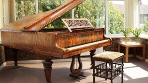 A grand piano in a sunlit room with large windows, a wooden bench, and a small table with a plant. - The Liver View (Wallasey)