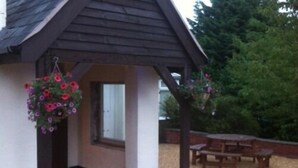 A small wooden structure with a bench and a table, surrounded by a gravel area and a tree. - The Liver View (Wallasey)