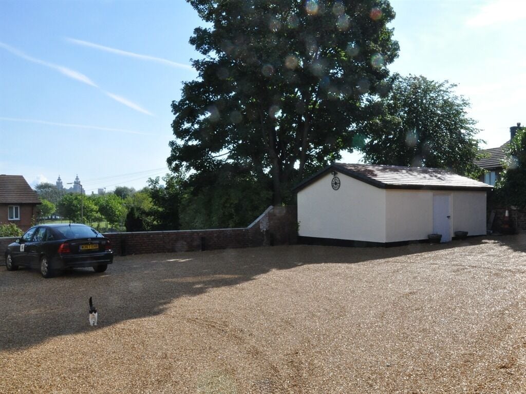 A gravel driveway leading to a white building with a sloped roof, surrounded by trees and a parked car.