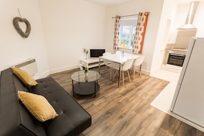 A modern living room with a black sofa, a round coffee table, and a white dining table with chairs. There is a TV on a stand, a small plant on the coffee table, and a kitchen area with a stove and oven. - Meridian Apartments - Southend (Southend-on-Sea)