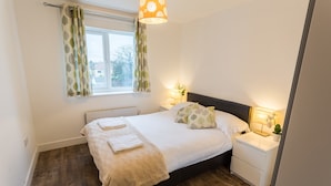 A bedroom with a bed, bedside tables, a window with curtains, a lamp, and a plant. - Meridian Apartments - Southend (Southend-on-Sea)