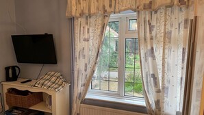 Un dormitorio con televisión, radiador, ventana con cortinas, cama y una pequeña mesa con tetera.