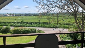 Vista desde un porche cubierto con baranda de madera, sobre un campo verde con algunos edificios a lo lejos.