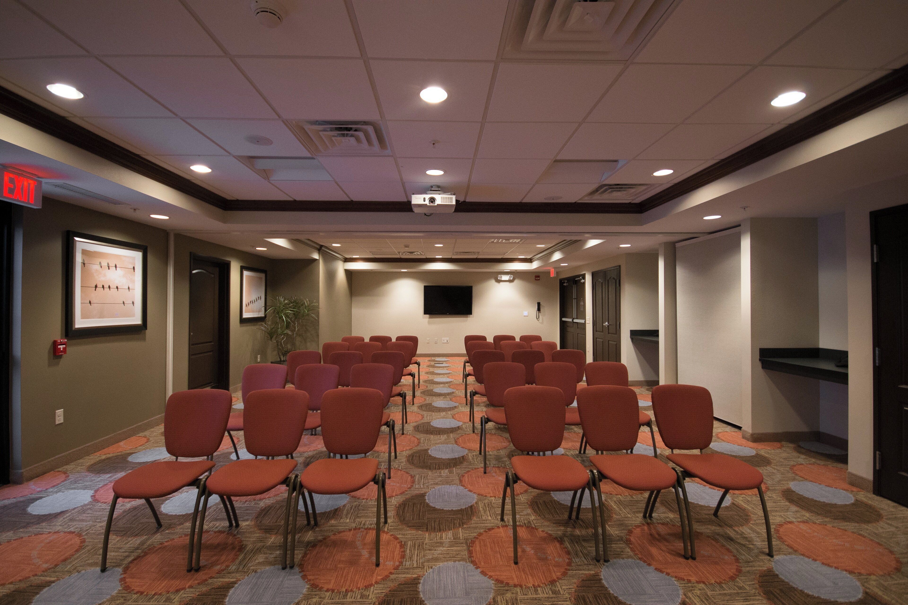 A conference room with rows of chairs, a projector screen, and a flat-screen TV on the wall.