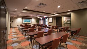 Uma sala de conferências com várias mesas e cadeiras, piso coberto de carpete e uma tela de projeção exibindo 'Staybridge Suites'.