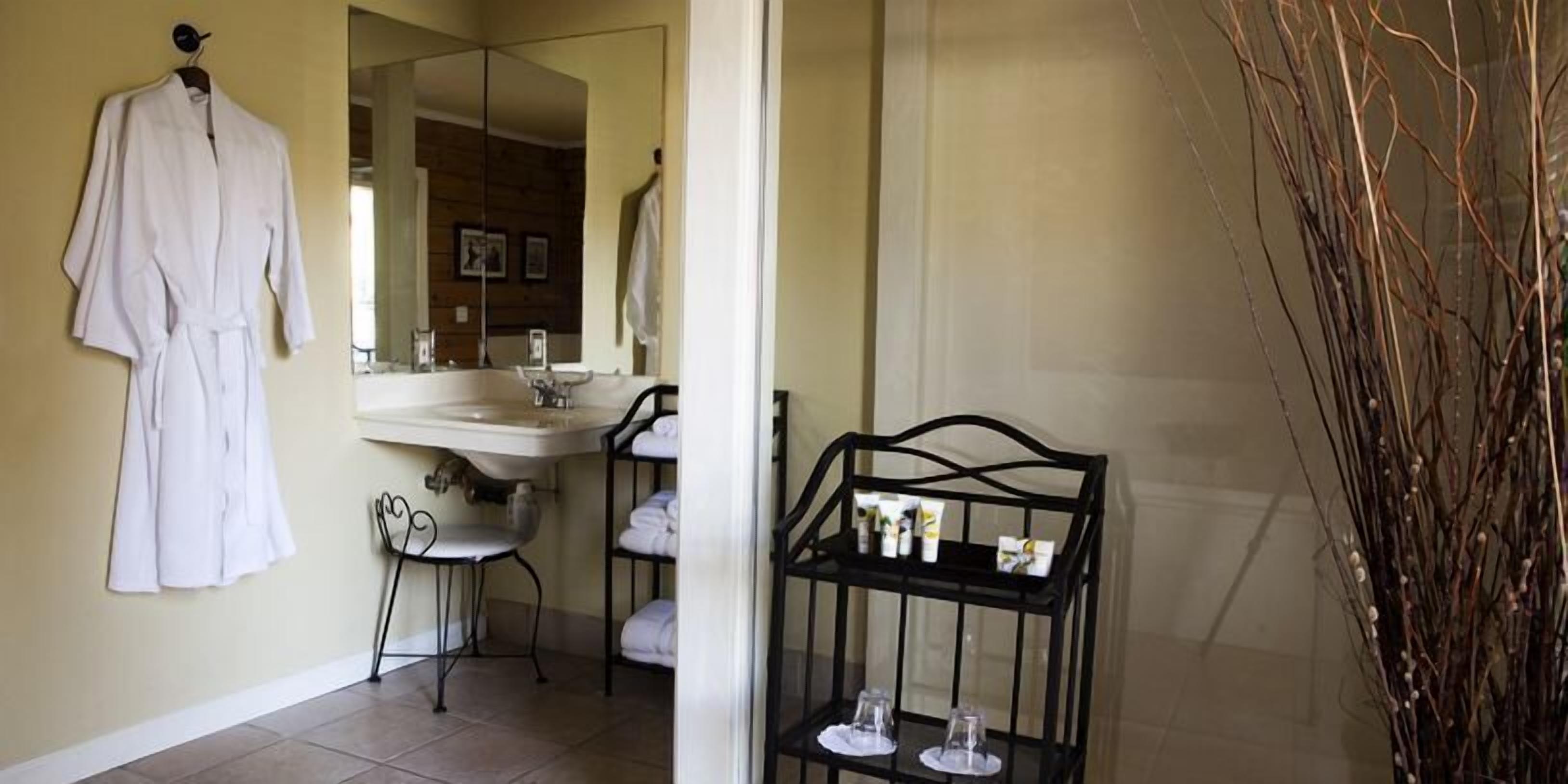 Un baño con bata blanca colgada, un lavabo, una silla y un estante con artículos de tocador.