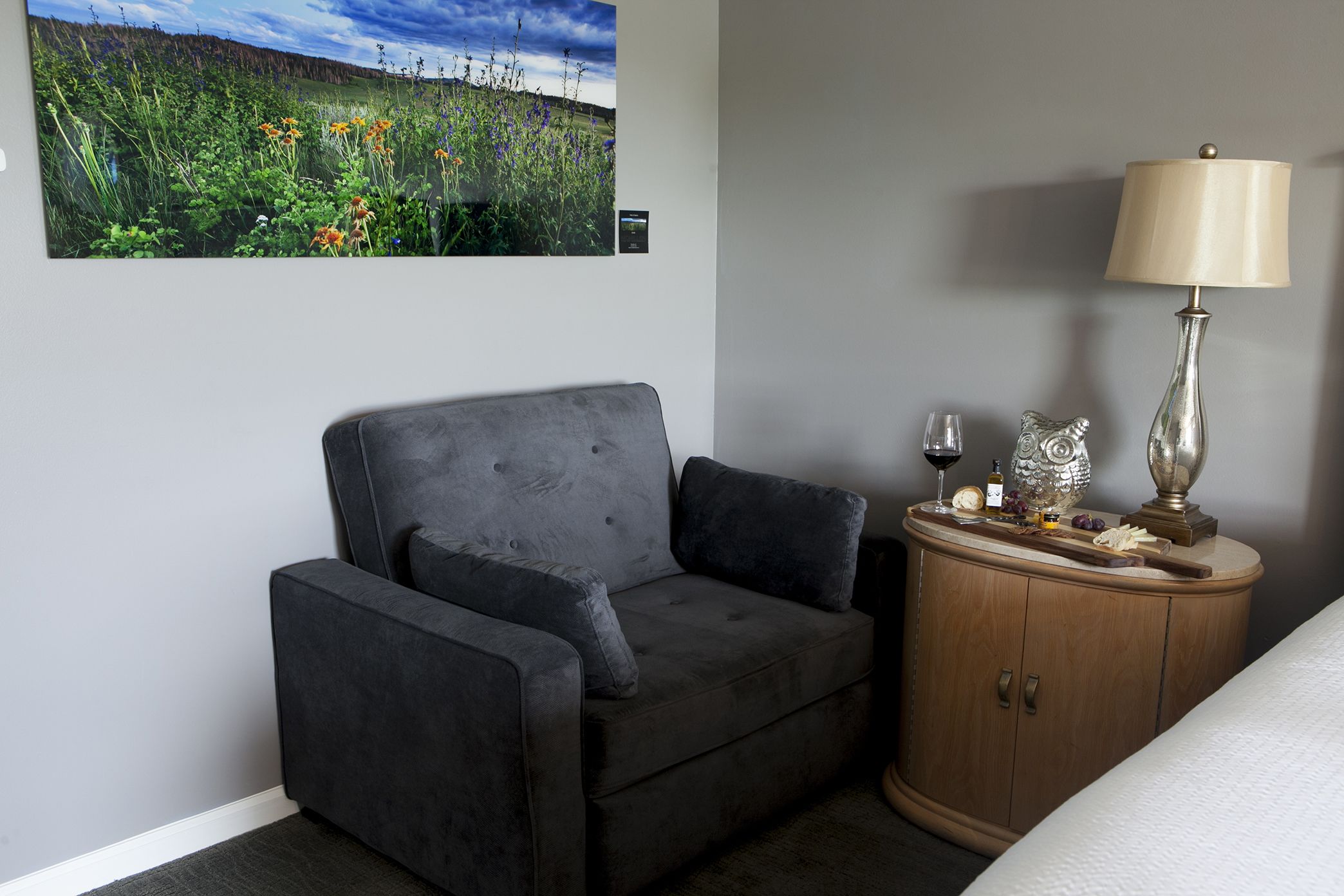 A room with a grey sofa, a wooden cabinet with a lamp, and a painting of a field.