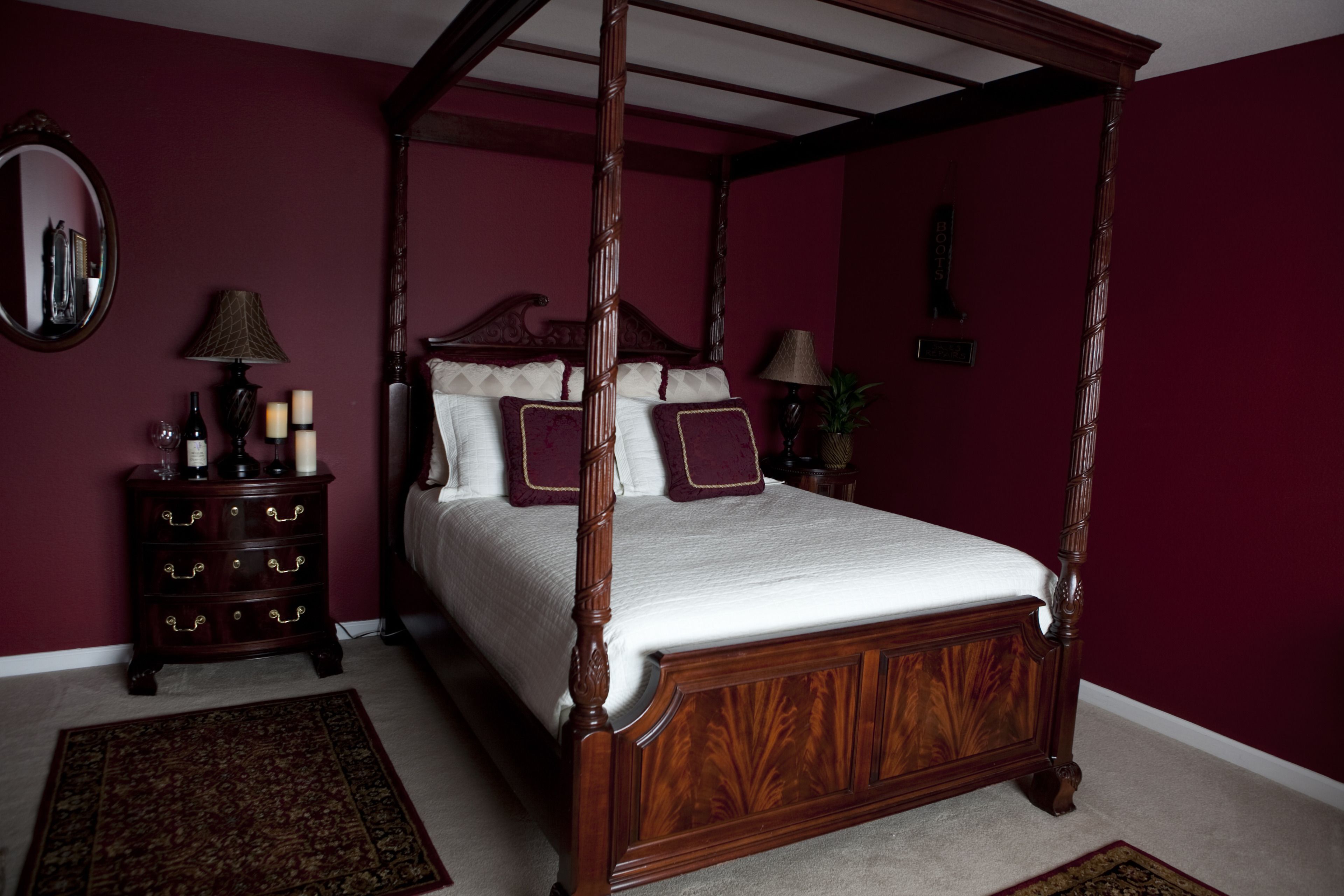 A four-poster bed with a wooden frame and white bedding, a dark wooden dresser with drawers, a round mirror, and a small table with a lamp and candles.