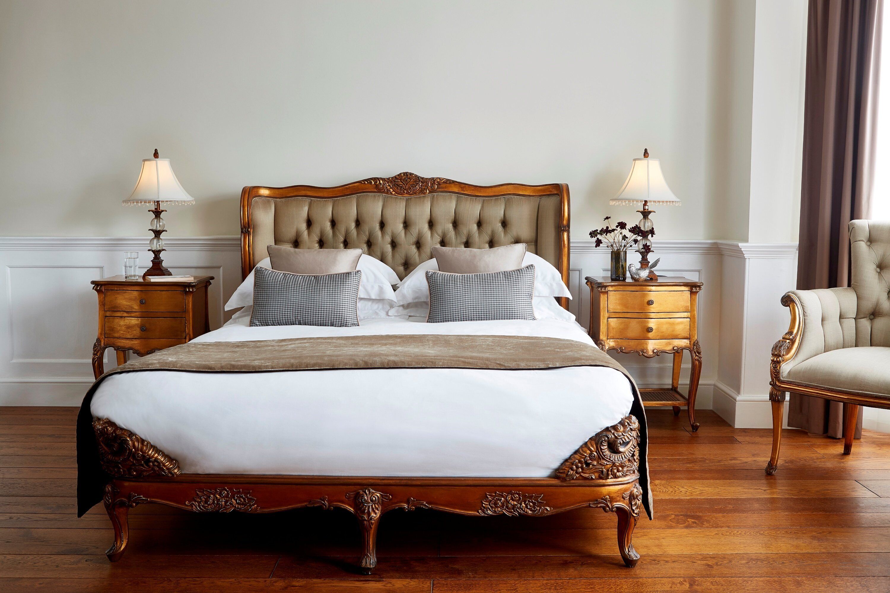 A large, ornate bed with a tufted headboard, flanked by two nightstands with lamps, in a room with wooden flooring and a classic chair.