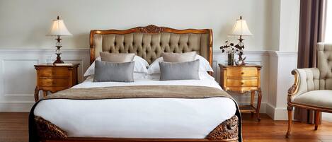 A large, ornate bed with a tufted headboard, flanked by two nightstands with lamps, in a room with wooden flooring and a classic chair.