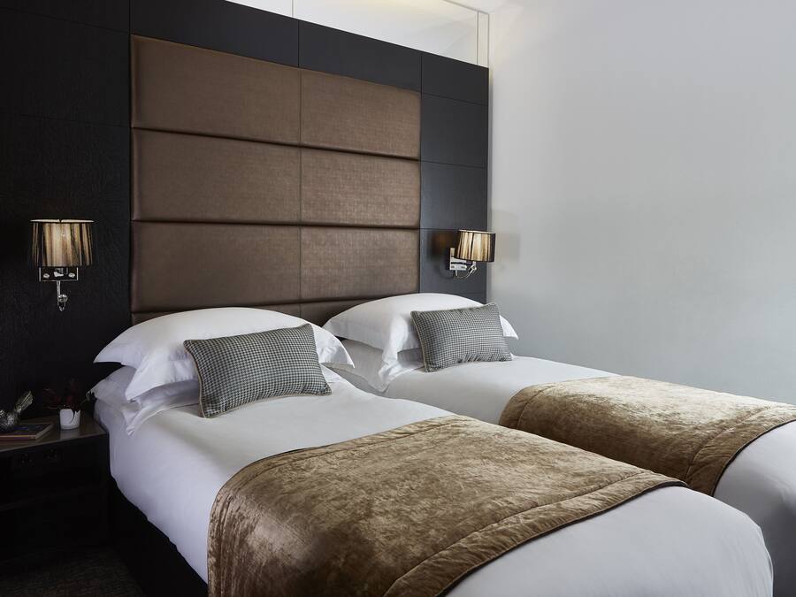 Habitación de hotel con dos camas, cabecero oscuro, lámparas de noche y mesita de noche con lámpara y un objeto decorativo.