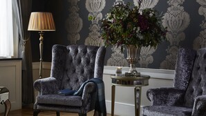 Una habitación con dos sillones de terciopelo, una mesita de noche con lámpara, y un jarrón con flores.