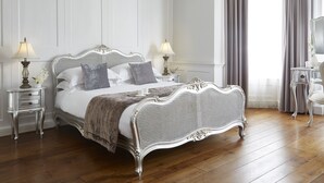 Una cama con dosel clásica, con detalles ornamentales, flanqueada por mesitas de noche con lámparas, en una habitación con suelo de madera y paredes blancas.