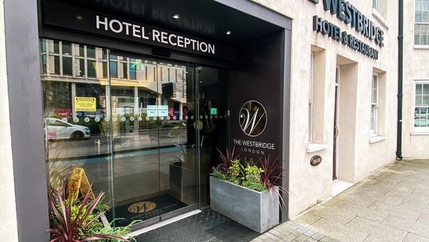 Recepción del Hotel Westbridge con entrada despejada, un cartel que indica que es hotel y restaurante, y dos macetas con plantas en el exterior.