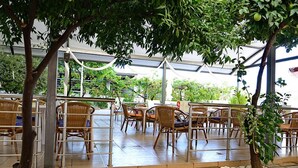 An outdoor seating area with wicker chairs and tables, surrounded by trees and a covered walkway. - Marianna Hotel (Kos)