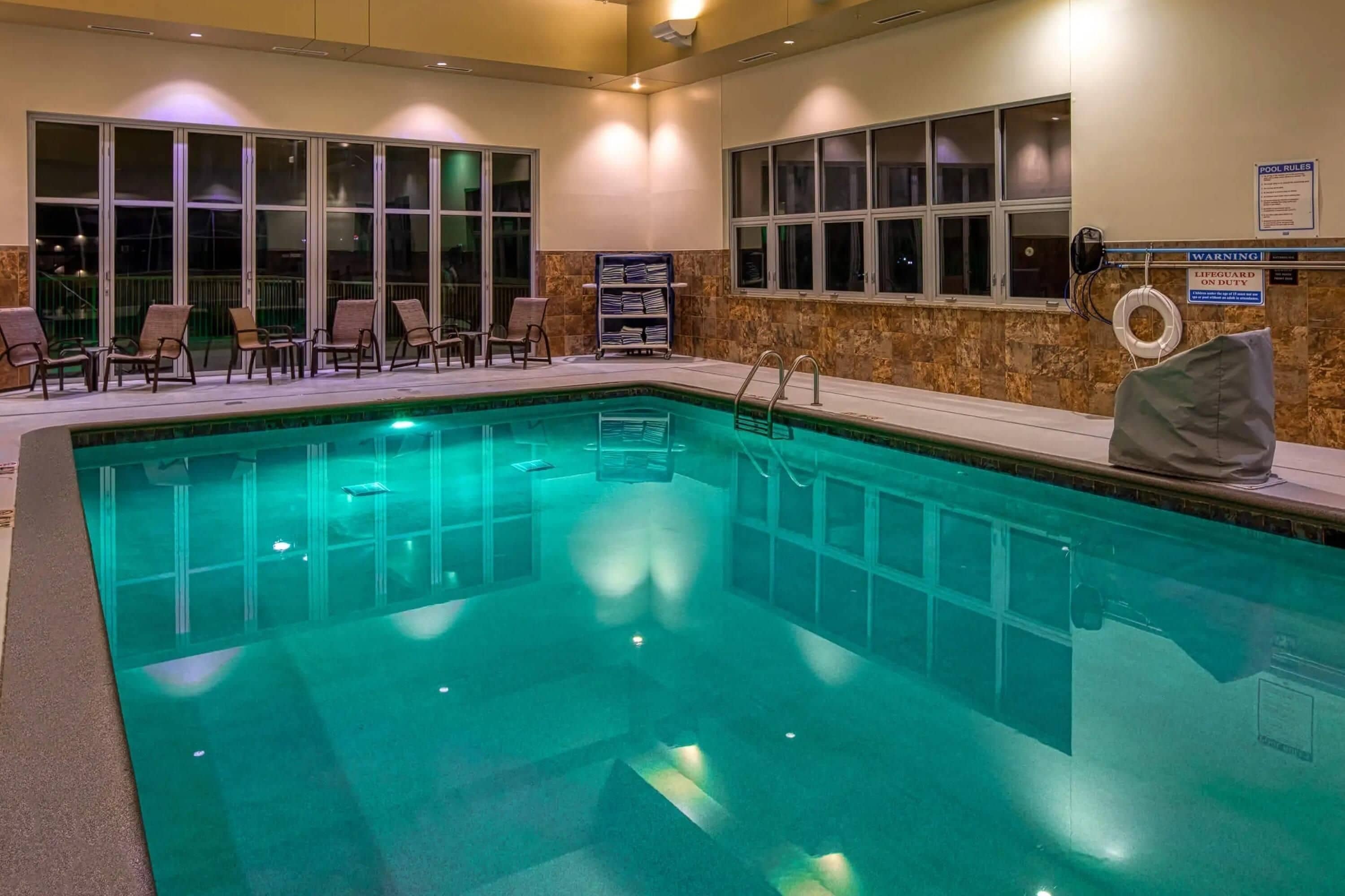 An indoor swimming pool with a lifeguard on duty sign, lounge chairs, and a pool rules sign.