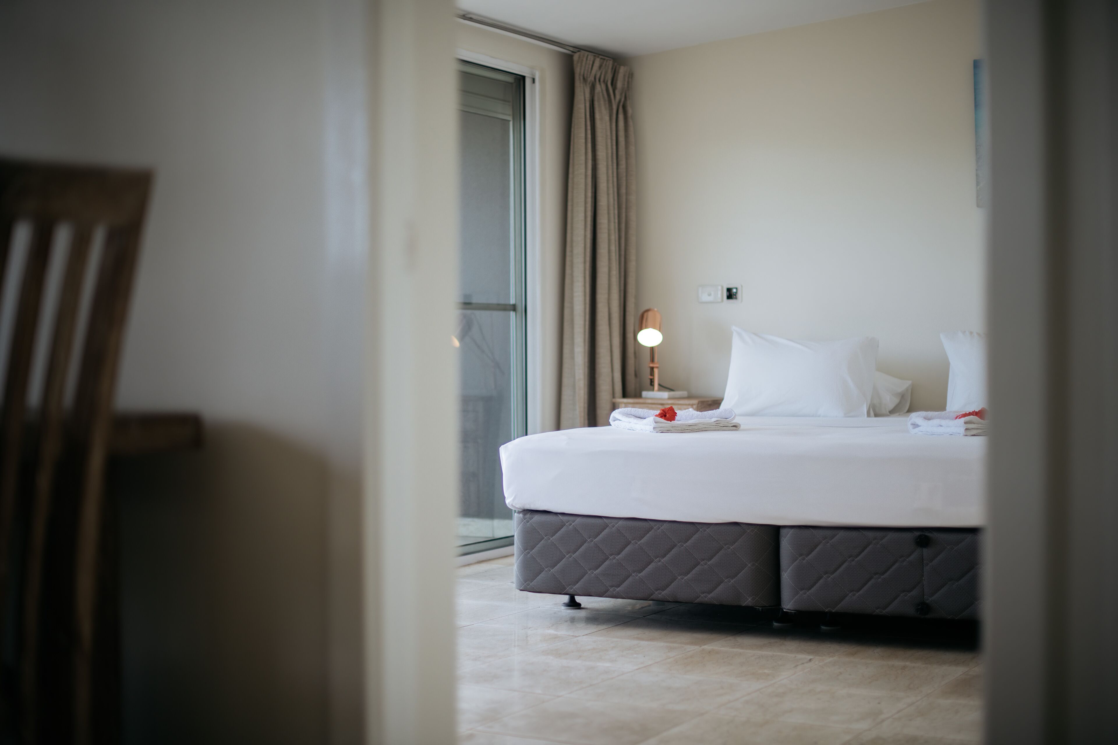 A hotel room with a bed, a nightstand, a lamp, and a view of the outside.