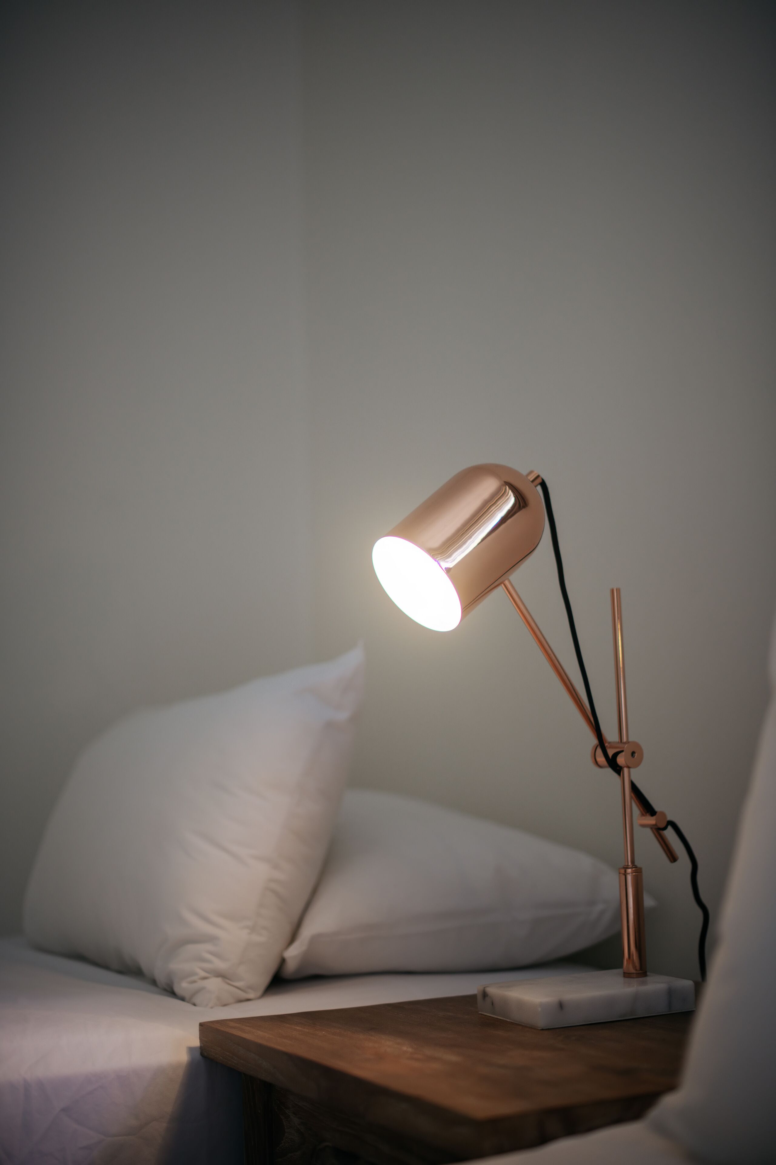 Ett sängbord med en modern lampa, en vit kudde och ett träbord som står bredvid sängen.