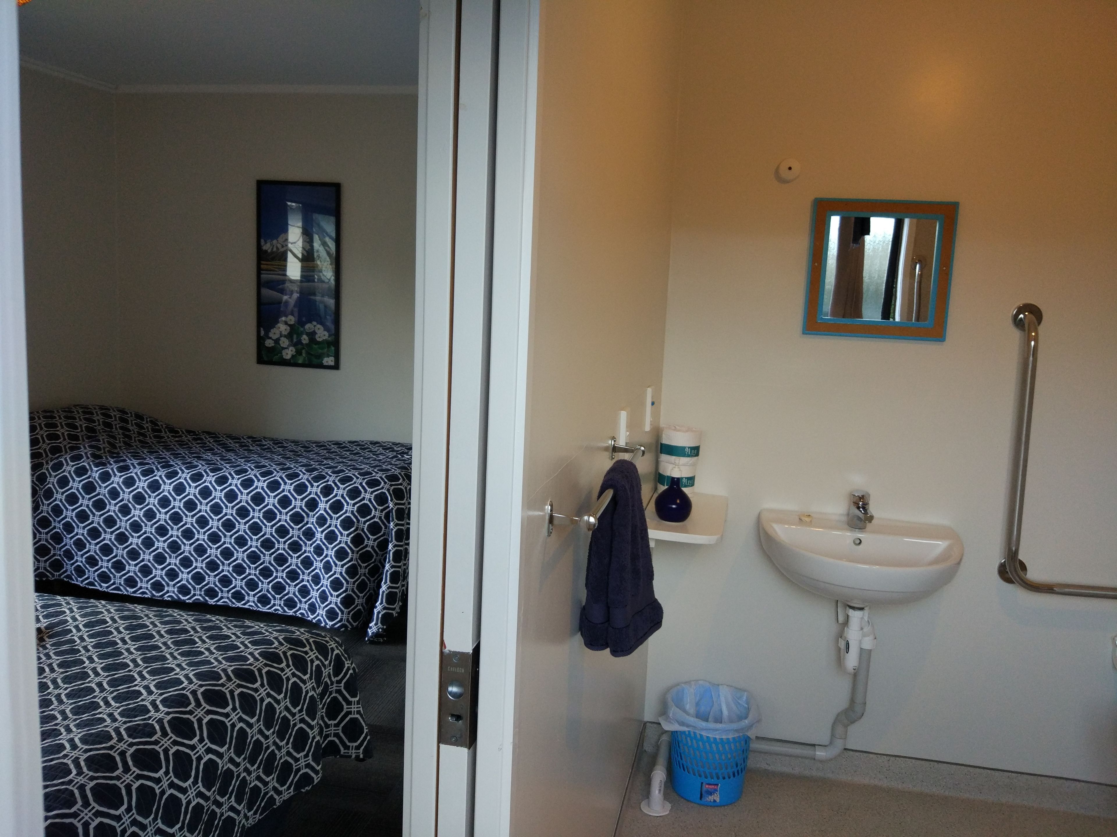 A bedroom with patterned bedding, a bedside table, a mirror, and a bathroom with a sink, toilet, and shower.
