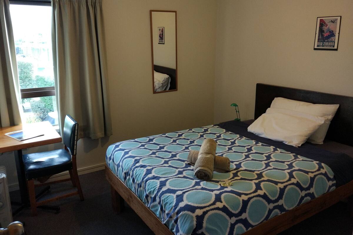 Une chambre d’hôtel avec un lit, un bureau, une chaise, une fenêtre avec des rideaux, une lampe de chevet et une affiche au mur.