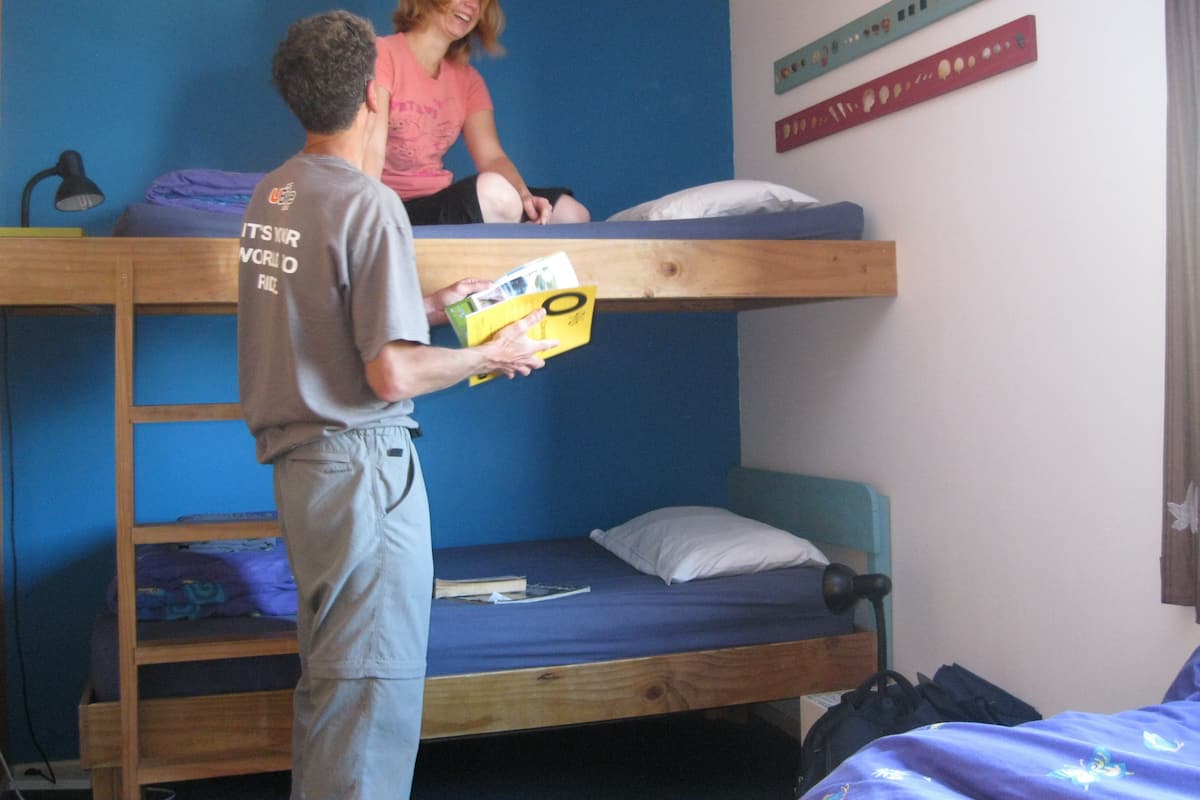 Une chambre avec un lit superposé. Une personne se tient debout sur le lit supérieur, un livre à la main, une autre personne est assise sur le lit supérieur, et il y a un bureau avec une lampe.