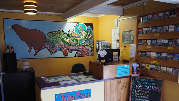 A reception desk with a map display, a large colorful mural, and a bookshelf with travel books. - Tasman Bay Backpackers (Nelson)