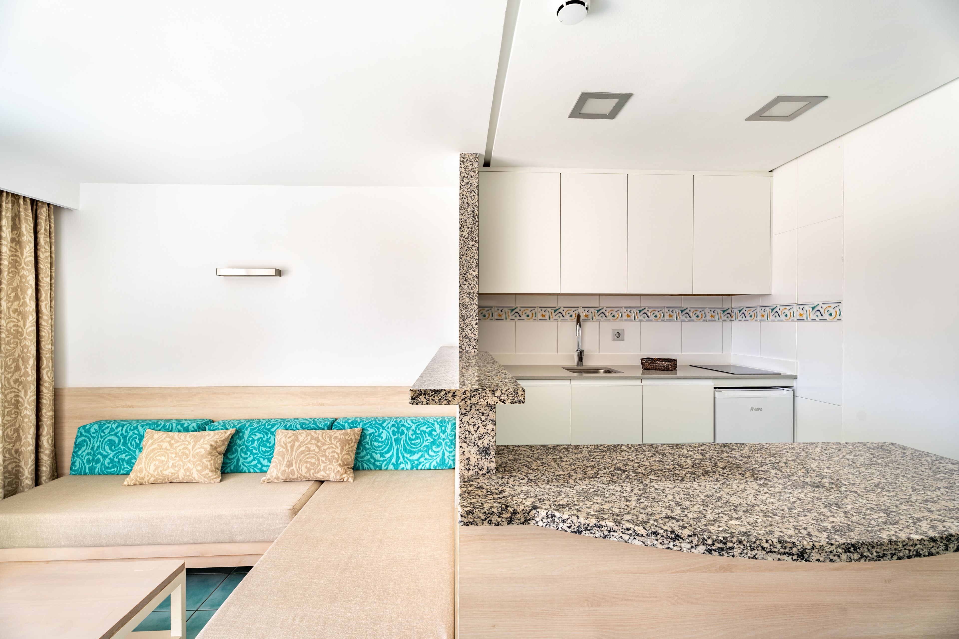 A modern, compact living space with a kitchenette, a sofa with patterned cushions, and a dining table.
