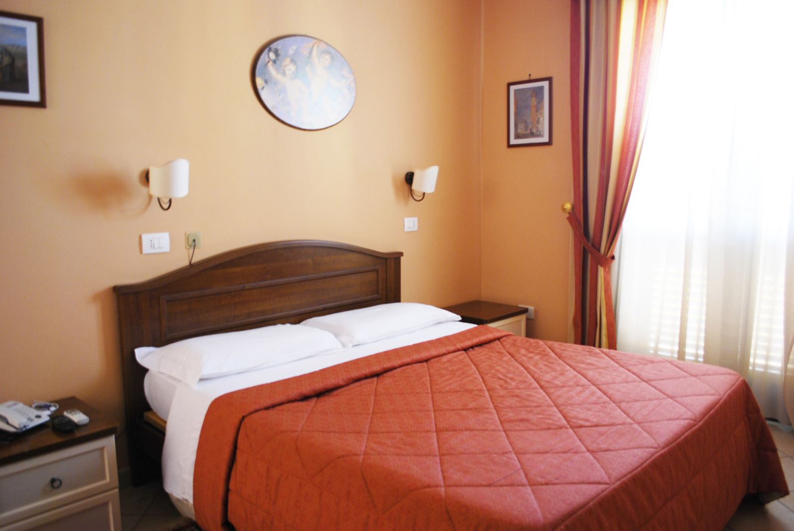 Quarto de hotel com uma cama grande, cabeceira de madeira, duas mesas de cabeceira, um criado-mudo e uma decoração redonda na parede, além de uma janela com cortinas.