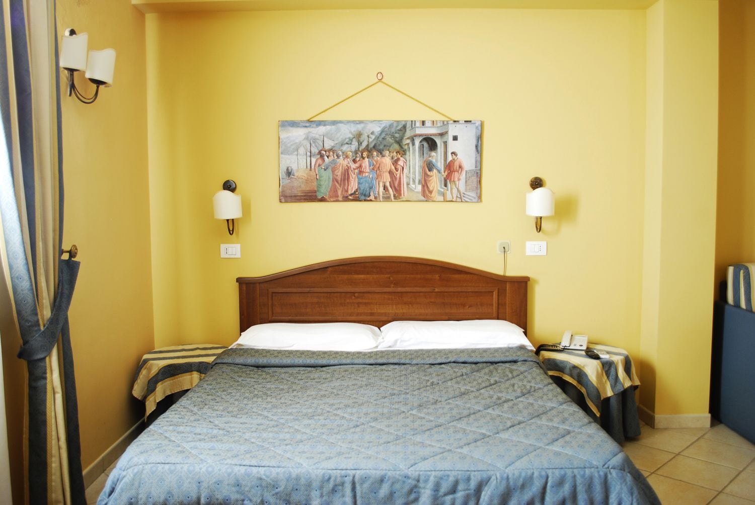 Habitación de hotel con una cama de madera, dos mesitas de noche, un cuadro en la pared y una cortina a la izquierda.