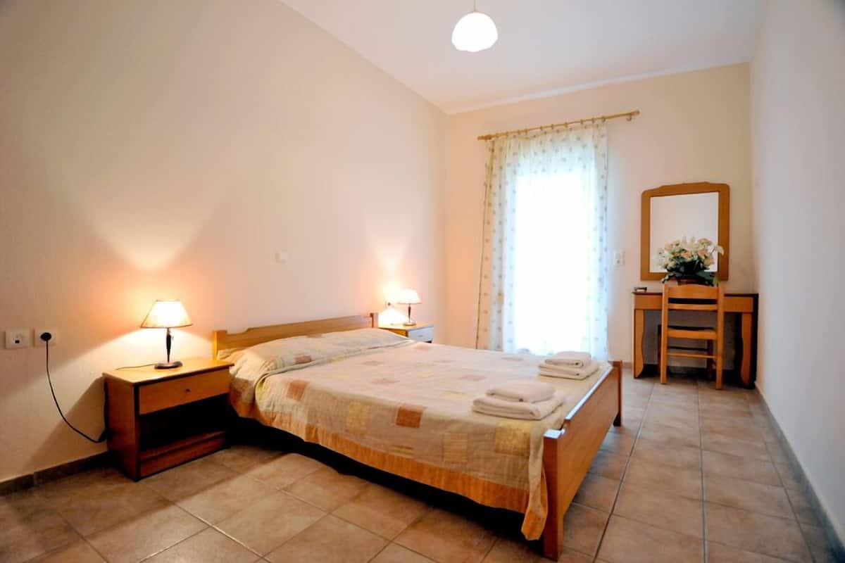 A bedroom with a bed, two bedside lamps, a wooden nightstand, a wooden chair, a mirror, and a window with curtains.